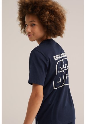 Jeune personne aux cheveux bouclés portant un T-shirt bleu marine avec le numéro blanc 56 et le texte "ESTABLISHED" dans le dos, regardant par-dessus son épaule.