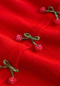 Červená texturovaná látka obsahuje dekorativní zelené korálkové mašle s růžovými korálkovými květinami, což vytváří detailní a složitý vzor.