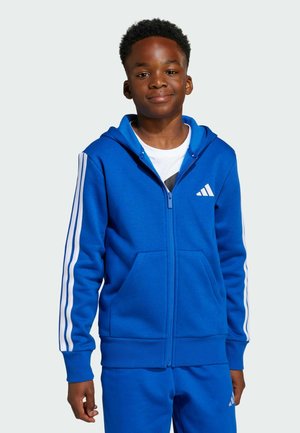 Chłopiec w niebieskim dresie Adidas z białymi paskami i logo, stojący na jasnym jednolitym tle, lekko się uśmiechając.