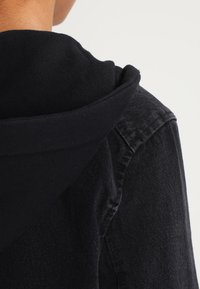 Musta huppari pehmeästä puuvillakankaasta, jossa on denim-yksityiskohtia. Ominaisuuksina pyöreä kaulus ja näkyvä sauma takana.