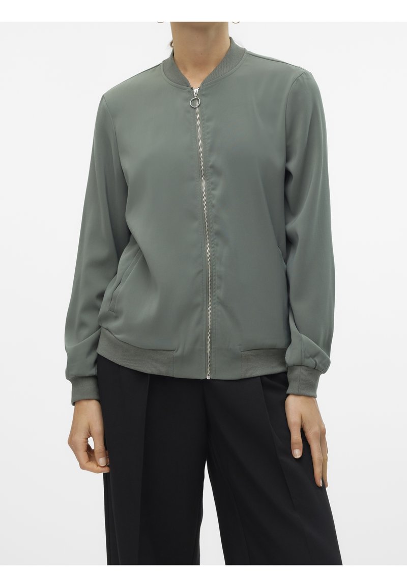 Femme portant une veste bomber zippée olive à manches longues avec poignets et bas en bord-côtes, associée à un pantalon noir à jambes larges.
