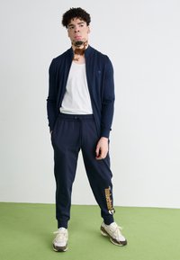 Chaqueta navy con cremallera y cuello alzado; camiseta blanca debajo; joggers navy a juego con detalle del logo en naranja; zapatillas gris y blancas.