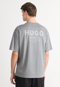 Grå bomulls t-skjorte med korte ermer, med en stor hvit "HUGO" logo og tekst på ryggen. Enkel, avslappet passform med rund hals.