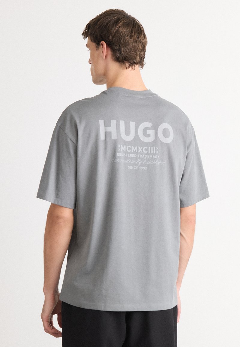 Grå bomulls t-skjorte med korte ermer, med en stor hvit "HUGO" logo og tekst på ryggen. Enkel, avslappet passform med rund hals.