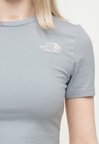 Κοντινό πλάνο ατόμου που φοράει ανοιχτό γκρι κοντομάνικο πουκάμισο με λευκό κεντημένο λογότυπο The North Face στο επάνω μέρος του στήθους.