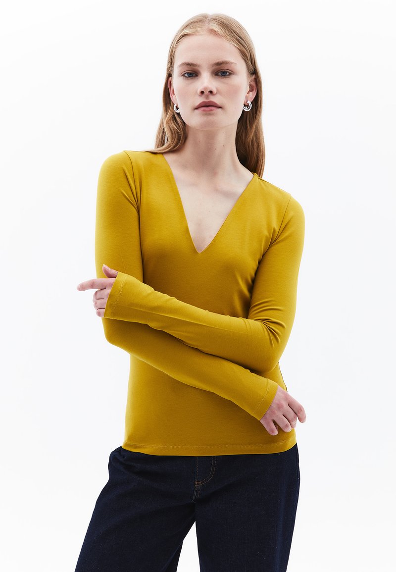 Maglietta gialla a maniche lunghe con scollatura a V profonda, tessuto morbido e silhouette aderente, abbinata a pantaloni in denim scuro.