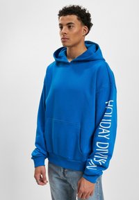 Sudadera con capucha azul, de mangas largas, con un bolsillo frontal y texto en blanco "HOLIDAY DIVISION" en la manga izquierda. Tejido suave, ajuste relajado.