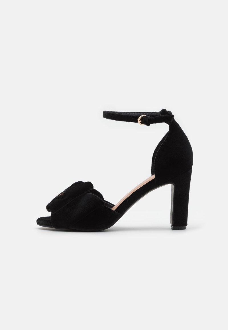 Sandale noire en daim à talon haut avec bride cheville et boucle, bout ouvert et détail nœud décoratif sur la bride avant.