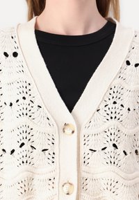 Strikket cardigan i lys kremfarge, med et blonde mønster, V-hals og to knapper foran. Båret over en svart ribbestrikket topp.