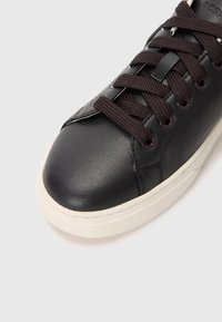 Svart lær sneakers med mørkebrune lisser, teksturert overflate og en hvit gummisåle. Rund tå og glatt overflate med subtile sømdetaljer.