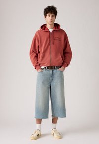 Sudadera roja con cremallera y texto "Two Horse Athletic Club", pantalones cortos de mezclilla anchos de color azul claro, y zapatillas de deporte beige con acentos oscuros.