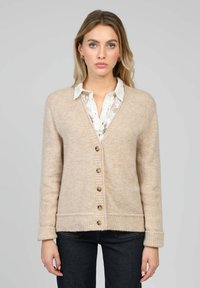 Cardigan beige en maille avec col en V, boutons en bois, poignets et ourlet côtelés, porté sur une chemise blanche à col en dentelle.