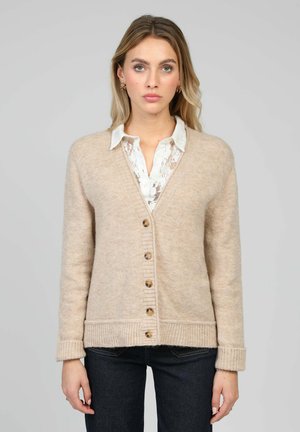 Cardigan beige en maille avec col en V, boutons en bois, poignets et ourlet côtelés, porté sur une chemise blanche à col en dentelle.