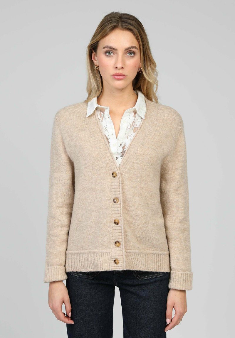 Cardigan beige en maille avec col en V, boutons en bois, poignets et ourlet côtelés, porté sur une chemise blanche à col en dentelle.