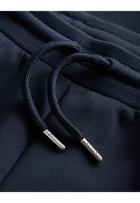 Marina träningsbyxor med strukturerade, flätade dragsnöre med silvriga metalldetaljer, mot en slät tygbakgrund.