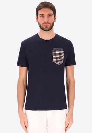 Uomo che indossa una t-shirt blu navy con una tasca sul petto a motivo pied-de-poule e pantaloni bianchi, in piedi davanti a uno sfondo uniforme.