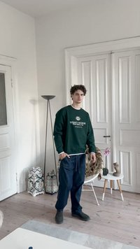 Zelená mikina s bielymi akcentmi a textom "LEGACY MANOR", v kombinácii s tmavomodrými nohavicami. Izba má biele steny, drevenú podlahu a dekoratívne akcenty.