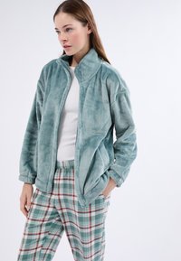 Kobieta w jasno niebieskiej polarowej kurtce, białej bluzce i zielono-czerwonych spodniach w kratę, stoi z jedną ręką w kieszeni na jednolitym tle.