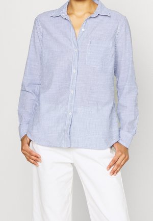 Kvinde iført en blå og hvidstribet skjorte med knapper og lange ærmer samt hvide bukser, stående med hænderne hvilende på hoften.