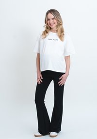Bílá bavlněná tričko s nápisem "COOL MAMA", spárované s černými rozšířenými kalhotami a bílými botami. Model stojí na neutrálním pozadí.