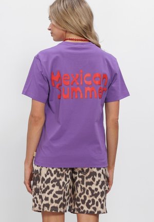 Vrouw met rug naar de camera, draagt een paarse T-shirt met rode tekst "Mexican Summer" en cargoshort met luipaardprint tegen een effen witte achtergrond.