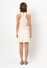 Witte kanten jurk met een tekstuurpatroon van bloemen, zonder mouwen en een sleutengatdetail op de rug. Kenmerkt zich door een franjeszoom en een getailleerd silhouet.