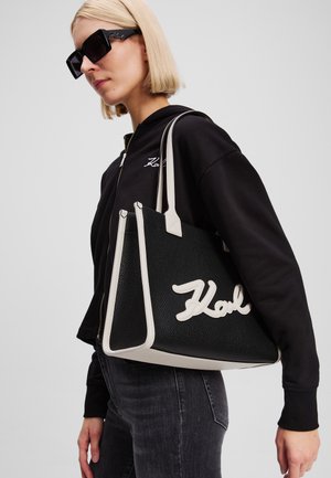Zwarte gevarieerde totebag met crèmekleurige accenten en logo. Heeft dikke straps en een hoekige vorm, gedragen door een persoon in casual kleding.