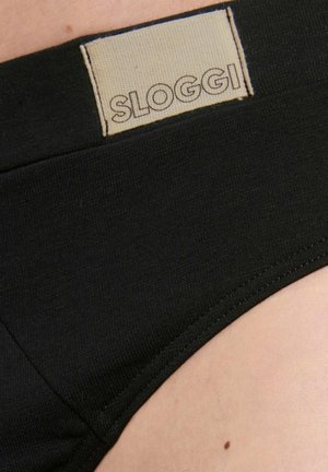 Nahaufnahme eines schwarzen Unterwäschebunds mit beigem rechteckigem Etikett, das "SLOGGI" in umrandeten Buchstaben auf der Haut zeigt.