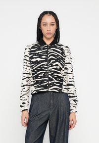 Veste zippée à manches longues avec rayures zébrées noires et blanches, col noir, portée sur un pantalon en denim foncé. Texture de tissu lisse.