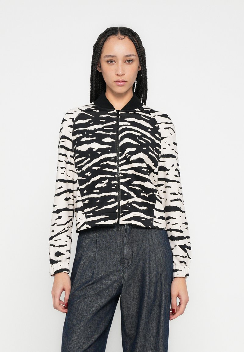 Veste zippée à manches longues avec rayures zébrées noires et blanches, col noir, portée sur un pantalon en denim foncé. Texture de tissu lisse.