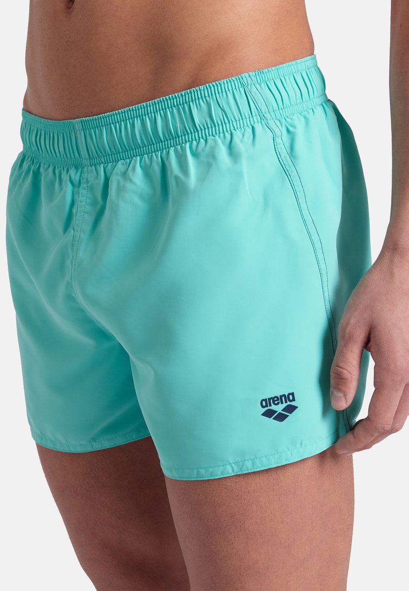 Mężczyzna w turkusowych kąpielówkach z elastycznym pasem i czarnym logo Arena na lewym udzie na jednolitym białym tle.