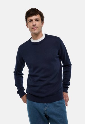 Pull en maille bleu marine avec un col rond, des poignets côtelés et une taille côtelée. Texture lisse, conçu pour un style décontracté, associé à un jean bleu clair.