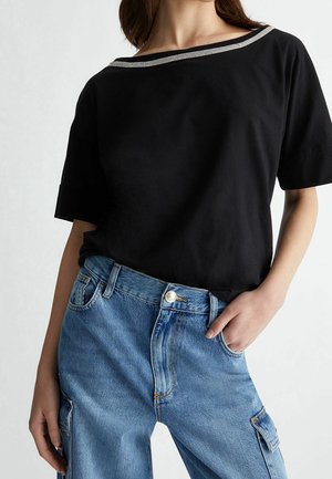 Femme portant un haut noir à manches courtes avec un col bordé d'argent et un jean cargo bleu, posant avec une main dans la poche.