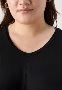Juodas marškinėliai su apvaliu iškirpimu, minkšta tekstūra ir trumpomis rankovėmis, akcentuojantys be siūlių dizainą iškirpimo vietoje.