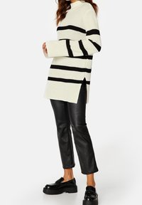 Gehaakte pullover in off-white met zwarte horizontale strepen, met een hoge halslijn, zijsplitten en lange mouwen, gecombineerd met zwarte leren broek.