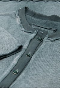 Gråblå stickad cardigan med rundad krage, fransade kanter och gröna knappar. Har en mjuk textur och en avslappnad design.