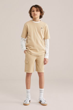 Jongen staand met handen in zakken, draagt beige cargoshorts, beige shirt met korte mouwen over een wit shirt met lange mouwen, gestreepte sokken en sneakers.
