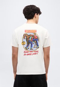 Άνδρας με σκούρα σγουρά μαλλιά που φοράει άσπρο T-shirt με πολύχρωμο vintage σχέδιο Levi Strauss με γραφικά αλόγου και κείμενο, που φαίνεται από πίσω.