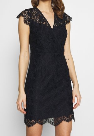 Frau trägt ein schwarzes Spitzenkleid mit kurzen Ärmeln, V-Ausschnitt und gezacktem Saum, zeigt ein kleines Tattoo auf ihrem linken Unterarm.