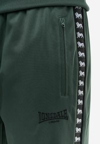 Dark green Lonsdale London pants with zippered pocket and black side stripe featuring white lion emblems along the seam.