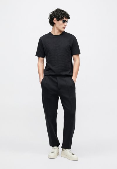 Čierna textúrovaná košeľa s krátkym rukávom kombinovaná s čiernymi upravenými nohavicami a krémovými teniskami. Nosené slnečné okuliare, ruky v vreckách.