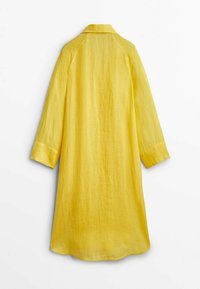 Cappotto giallo in lino con colletto, maniche lunghe a raglan e taglio dritto. Il tessuto ha un aspetto leggero e texture.