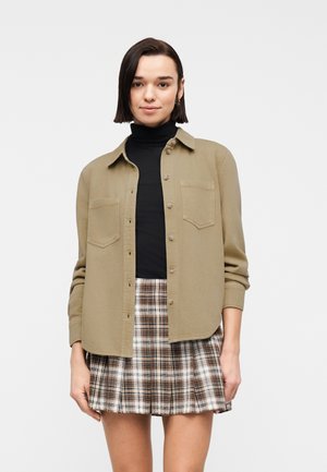 Young woman with short dark hair wears a black turtleneck, beige buttoned shirt, and pleated brown and white plaid skirt against white background.