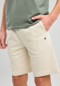 Pantalones cortos de algodón beige con bolsillos, con un pequeño logo bordado en azul, rojo y blanco. Corte recto y dobladillos cosidos.