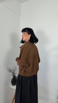 Giacca oversize in suede marrone con orlo a coste, abbinata a una lunga gonna nera. Texture liscia e vestibilità rilassata. Elementi di sfondo minimalisti.