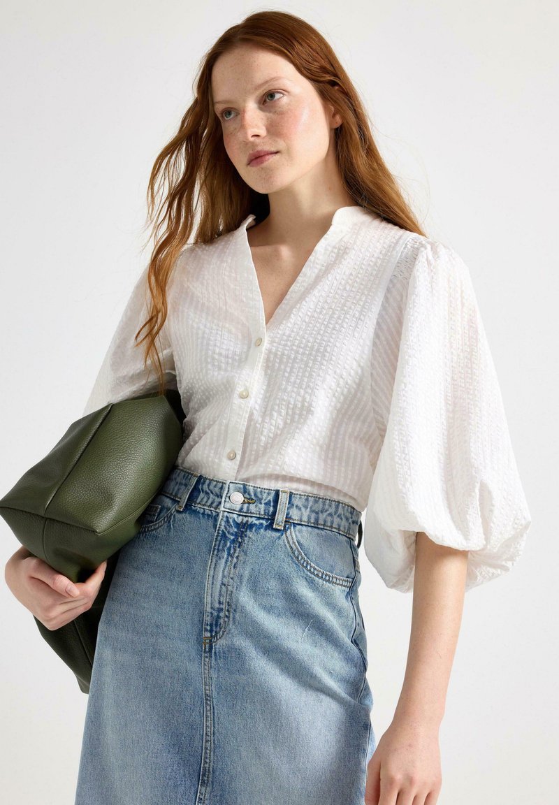 Vit texturerad blus med knappstängning, med puffärmar. Kombinerad med en högmidjad denimkjol och en grön läderväska.