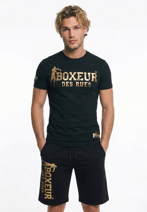 Blonder Mann trägt schwarzes T-Shirt und Shorts, beide mit goldenem Schriftzug „BOXEUR DES RUES“ und Boxfigur, steht mit den Händen in den Taschen.