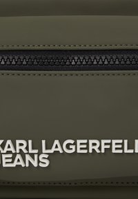 Ελιά πράσινη θήκη με λεία υφή, που διαθέτει μαύρο φερμουάρ και έντονο άσπρο κείμενο που λέει "KARL LAGERFELD JEANS."