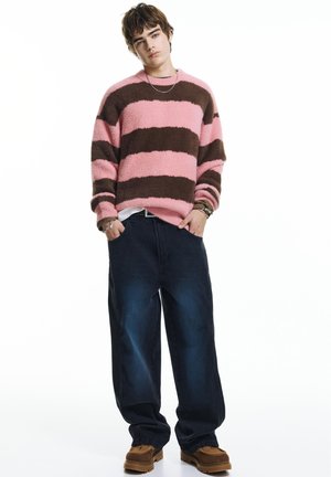 Jeune homme portant un pull duveteux rayé rose et marron, un jean foncé surdimensionné et des chaussures marron, debout les mains dans les poches.