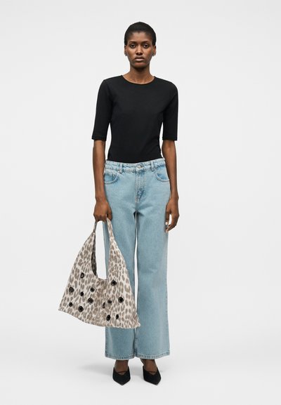 Γυναίκα που φοράει μαύρο τοπ και φαρδιά παντελόνια τζιν ανοιχτού γαλάζιου χρώματος, κρατώντας μια μεγάλη μπεζ τσάντα με λεοπάρ σχέδιο και μαύρες τρύπες.